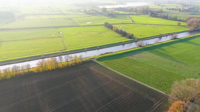 Aerial view of rural fields divided by hedgerows, canal, and scattered trees sunlit landscape with water patches, paths, and distant farmhouses in vibrant countryside.