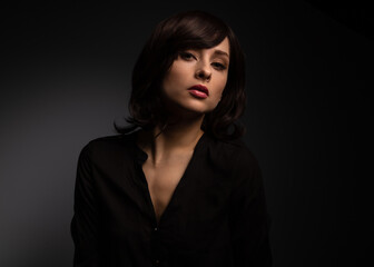 Fototapeta premium Sexy makeup female model with black short hair posing in black cotton elegant shirt on dark shadow background with empty space. Closeup fashion style portrait