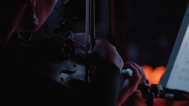 A close-up of a violinist's hand skillfully playing the violin, against a dark background illuminated by a soft orange light. The musician demonstrates the pizzicato technique