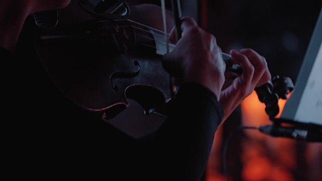 A close-up of a violinist's hands skillfully playing violin using the pizzicato technique, against a dark background illuminated by the soft orange glow of candles. A string concert by candlelight
