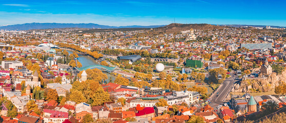 Panoramic view of Tbilisi, Georgia, in autumn, showing the Kura river and Rike Park under a blue sky © EdNurg
