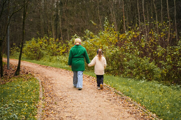 Obraz premium Walking mother with daughter on park path. Holding hands shows parenting, care, trust, safety. Outdoor family activity supports bonding, happiness, healthy lifestyle, weekend leisure, forest trail.