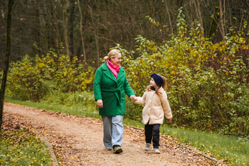 Fototapeta premium Walking mother with daughter on park path. Holding hands shows parenting, care, trust, safety. Outdoor family activity supports bonding, happiness, healthy lifestyle, weekend leisure, forest trail.