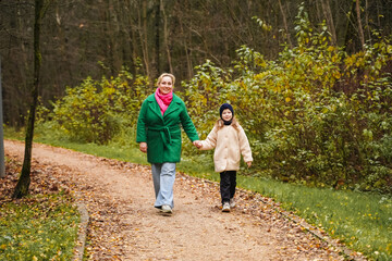 Fototapeta premium Walking mother with daughter on park path. Holding hands shows parenting, care, trust, safety. Outdoor family activity supports bonding, happiness, healthy lifestyle, weekend leisure, forest trail.