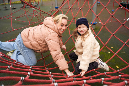 Balancing mother with child on rope playground net. Climbing swing game builds trust, strength, coordination. Outdoor family play supports fitness, courage, teamwork, weekend leisure, happy childhood. - Powered by Adobe