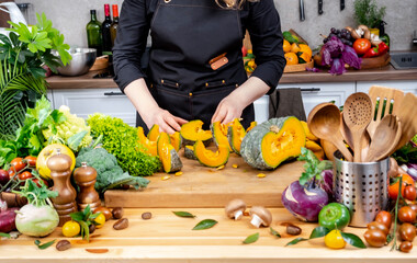 Cook arranging pumpkin slices on wooden board with fresh vegetables in bright home kitchen, healthy...