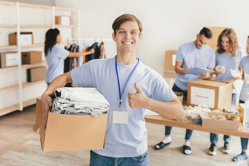 A group of volunteers work together in a community center, sorting clothing donations. One volunteer smiles and holds a box of clothes while others organize items at tables in the background.