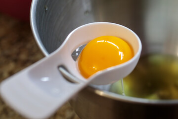 Close-up of a bright, intact egg yolk being held in a white plastic separator spoon over a stainless steel mixing bowl with a stand mixer whisk visible