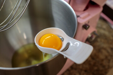 Close-up of a bright, intact egg yolk being held in a white plastic separator spoon over a stainless steel mixing bowl with a stand mixer whisk visible