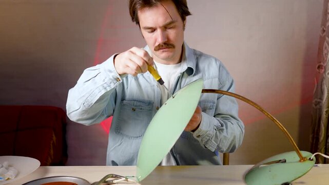 Craftsman restorer master working on refurbishing used vintage electric lamp at home workshop, man screwing in big lampshade to original lamp base with screwdriver tool, checking item conditions.