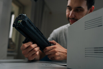 Close-up portrait of bearded repairman smiling performing printer maintenance, replacing empty toner cartridge with new one on desk, ensuring smooth office operations and diy repair.