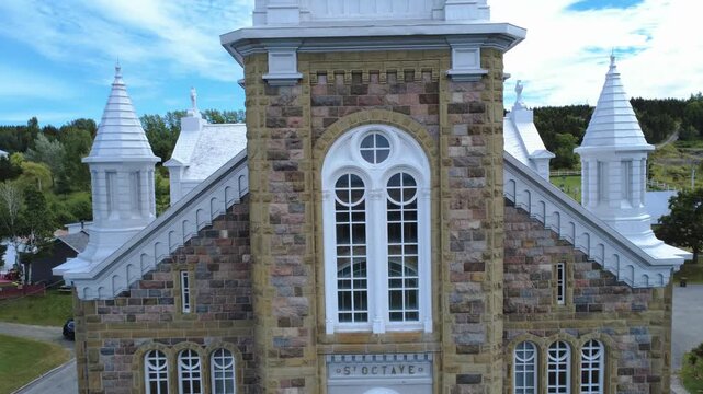 Upward view of the brick and stone facade of the Church of Saint-Octave-de-M&eacute;tis with its large windows and its bell tower with three bells up to its spire surmounted by a cross-shaped lightning rod.