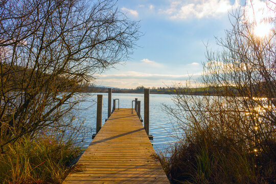 Serene wooden pier extending into calm lake under dramatic sky, surrounded by bare trees. Peaceful nature scene evoking relaxation, contemplation and quiet winter atmosphere. - Powered by Adobe