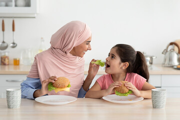 A mother and her young daughter share quality time while enjoying burgers at a cozy kitchen table. They smile and interact happily during their meal.