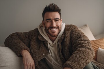 Brightly lit living room with soft natural light. Handsome Latin man in casual attire sits relaxed on a plush sofa, smiling warmly at the camera