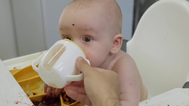Baby with a messy face sits in a high chair and happily with appetite drink water from a sippy cup. Concept of early childhood, feeding time, and introducing solids to infants. parental care