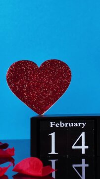 Romantic concept for Saint Valentine's Day with red rose petals falling in slow motion over a perpetual calendar showing February 14th and a glittering heart against a vibrant blue background