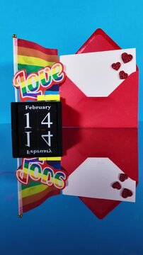 Slow motion footage showing a Valentine's Day composition with a red envelope, a perpetual calendar marking February 14, and an lgbtq rainbow flag, all reflected on a mirror surface