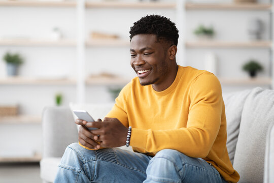 Handsome black guy in casual sitting on couch, chatting with girlfriend, making online order via mobile application, using cellphone at home, copy space. Modern technologies and millennials lifestyle