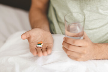 A person sits on a bed, holding a few colorful pills in one hand and a glass of water in the other. The scene suggests a moment of self-care and health management during the day.