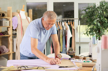 Mature male tailor stands near a table in the workshop and makes markings for patterns on paper....