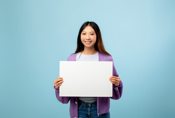 Fototapeta premium A young woman stands against a bright blue background, holding a blank white sign. She smiles warmly while wearing a purple cardigan and casual jeans.