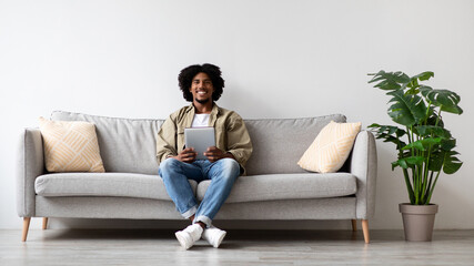 A young African American man is comfortably seated on a modern couch. He smiles while holding a digital tablet, enjoying his leisure time in a bright living room with plants.