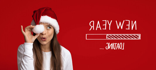 A young woman joyfully wears a Santa hat and playfully poses against a bright red background. She appears to be anticipating the New Year, creating a festive atmosphere.