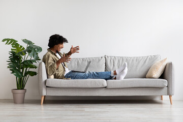 A joyful young African American man makes a video call from his living room. He sits comfortably on a couch, raising his hands in excitement during the online conversation.