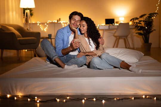 Millennial spouses are sitting together on a mattress, expressing love and affection while enjoying a relaxed evening at home. Their cozy bedroom is softly lit with warm lights.