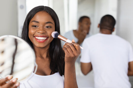 A woman with long dark hair smiles as she applies blush in front of a mirror. A man in a white shirt is visible in the background, completing his grooming.