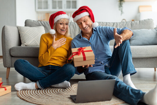 A joyful couple wearing Santa hats sits on the floor of their living room. They share laughter while opening a gift during a video call, surrounded by holiday decorations. - Powered by Adobe