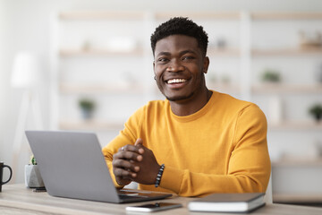 Portrait of positive young black guy in yellow sweater sitting at table, working on laptop, sending...