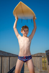 Smiling boy in swim trunks holding his surfboard high over his head on a boardwalk near the beach