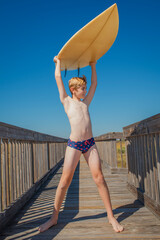 Smiling boy in swim suit carrying his surfboard over his head on a walkway near the beach