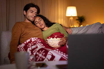A couple is sitting on a couch wrapped in a cozy blanket, watching Christmas movies on a laptop. They hold a bowl of popcorn while enjoying a quiet night together at home.