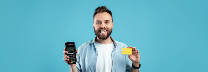 A young man with a beard happily holds a payment terminal in one hand and a credit card in the...