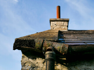 A roof with a chimney and a damaged water drain pipe. The roof is old and the pipe is rusty and vintage. Old stone building.