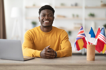Happy african american man teacher working at language school, sitting at workdesk with notebook and flags of different countries, looking at camera and smiling, office interior, copy space © Prostock-studio