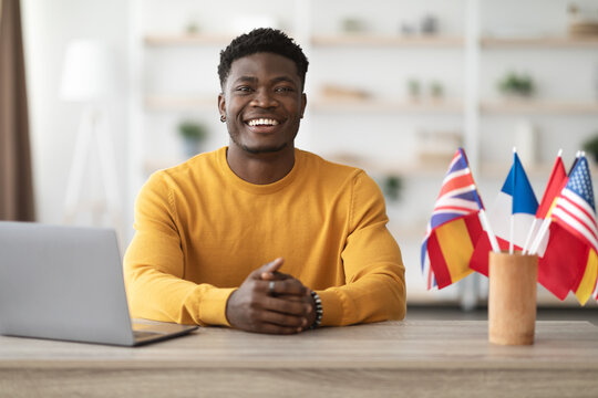 Happy african american man teacher working at language school, sitting at workdesk with notebook and flags of different countries, looking at camera and smiling, office interior, copy space