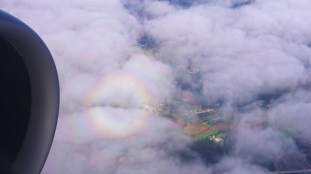 Daytime aerial over Madeira near Funchal shows jet engine at left, soft clouds, farmland glimpses, and a circular glory around the plane shadow on cloud tops.