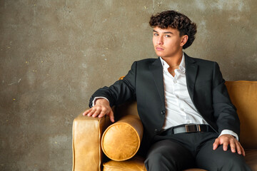 Studio portrait of a smiling teenage male smartly dressed in a prom suit casually sitting on a modern leather couch © 3Days2Go Media