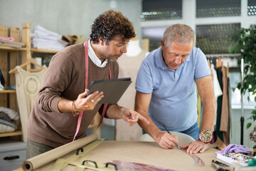 Adult male tailor showing elderly male assistant pattern drawing on tablet in sewing workshop