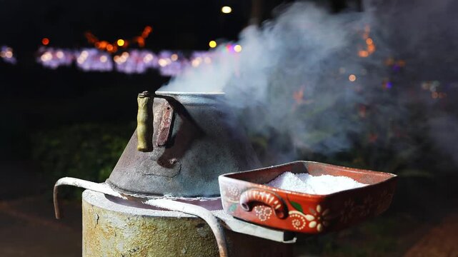 A rusted conical stove holds a painted pan of coarse white crystals as smoke drifts right in Funchal, Madeira at night, with festive lights and shallow depth of field.
