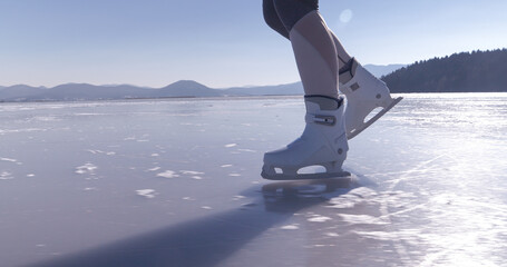 CLOSE UP: Woman in white ice skates glides across the smooth frozen surface of scenic Lake Cerknica in Slovenia. Ice skating on a cold and sunny winter day with a view distant hills in the background.