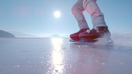 LOW ANGLE VIEW, LENS FLARE, CLOSE UP: Man in black ice skates brakes on a glossy ice surface of frozen Lake Cerknica as sparkling white ice crystals fly towards camera. Fun outdoor activity in winter. © helivideo