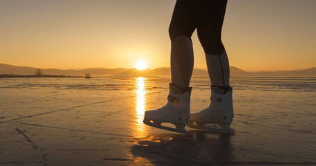 LENS FLARE, CLOSE UP, SILHOUETTE: Woman with white ice skates glides across frozen Lake Cerknica at sunset. Soft evening light of setting winter sun creates golden reflections on smooth icy surface.