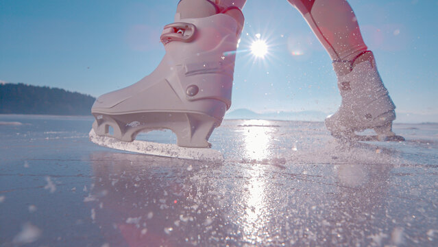 CLOSE UP, LENS FLARE, LOW ANGLE VIEW, DOF: White ice skates and blades on smooth surface of a frozen lake at sunset. Unknown woman figure skating and spraying white ice crystals while making a turn.