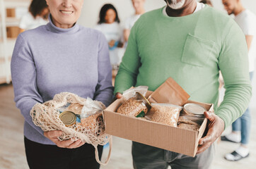 A man and a woman hold boxes of food items while participating in a community charity event focused on providing essential supplies. Others can be seen volunteering in the background.
