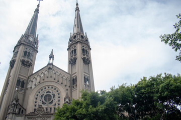 Majestic gothic church façade with tall twin towers, intricate architectural details, and a large rose window rising above lush green trees under a bright cloudy sky